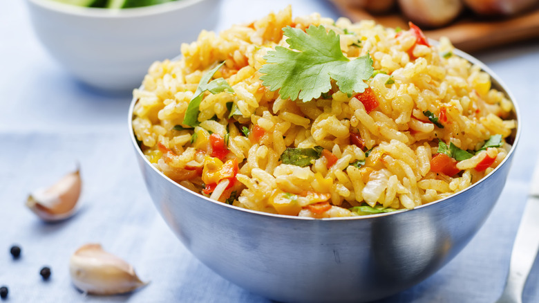 A stainless steel bowl of saffron rice with herbs and vegetables topped with a cilantro leaf on a blue tablecloth, surrounded by other bowls, two garlic cloves and three black peppercorns.
