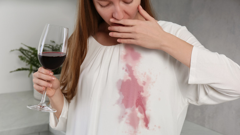 A woman looking down at a large red wine stain on her white blouse.