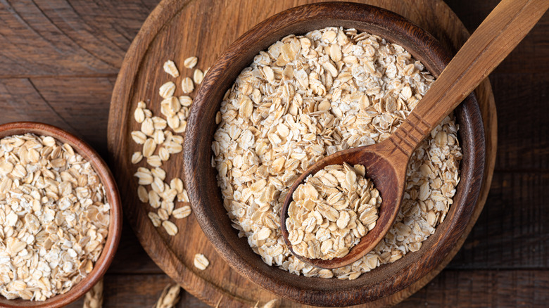 Raw rolled oats in wooden bowl on top of cutting board with wooden spoon next to smaller dish of oats.