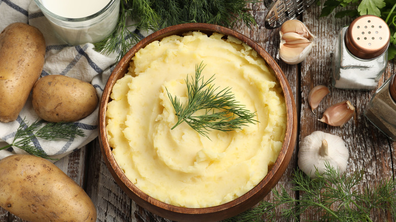 Bowl of mashed potatoes on a wooden table with salt, garlic, herbs, and raw potatoes