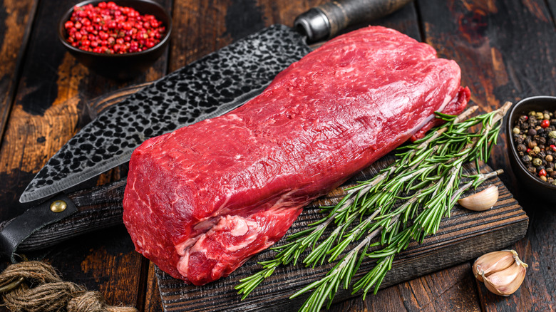 Whole beef loin on a wooden cutting board with herbs, spices, and a knife