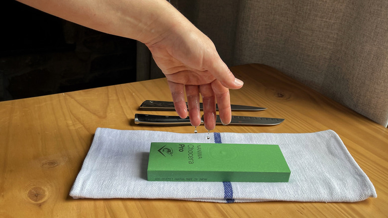 A hand drips water onto a whetstone over a towel with knives in the background