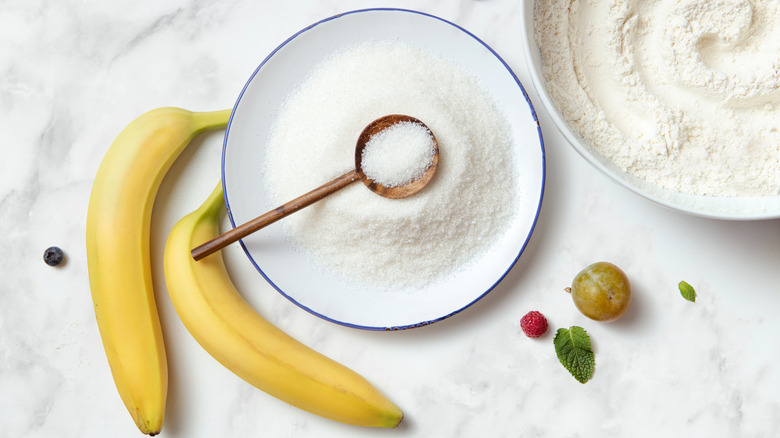 White sugar in a bowl and bananas on a white countertop.