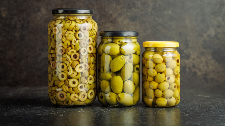 Multiple jars of pickled green olives in brine on a black table.