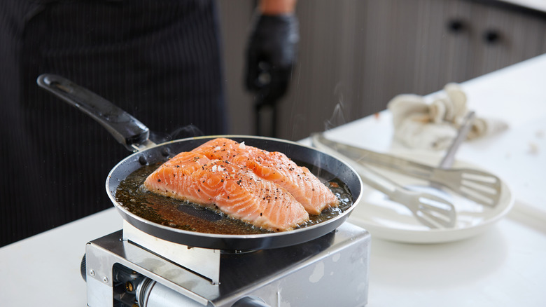 salmon dimasak dalam wajan dengan koki di latar belakang