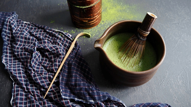 Brewed matcha tea in a brown bowl with a whisk and a tea towel on the side.