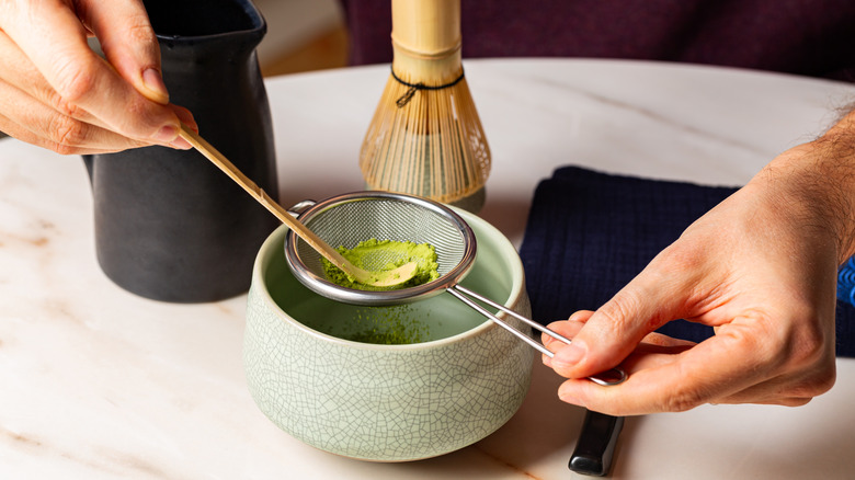 An individual sifting green matcha tea through a sieve and into a bowl.