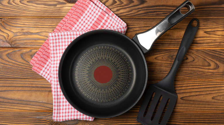A nonstick pan placed on a wooden surface, along with a red cloth and a black plastic spatula