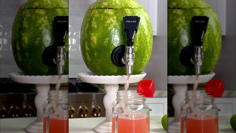 A watermelon with a drink spout sitting on a cake stand and dispensing a fruity drink into a Mason jar glass with a handle