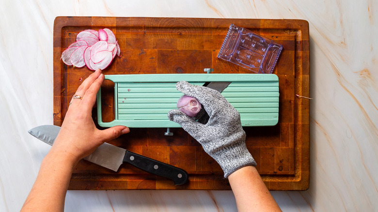 Someone wearing a glove on one hand and cutting a shallot on mandoline on top of a cutting board