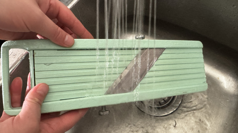 A person washing a mandoline under running water in a sink
