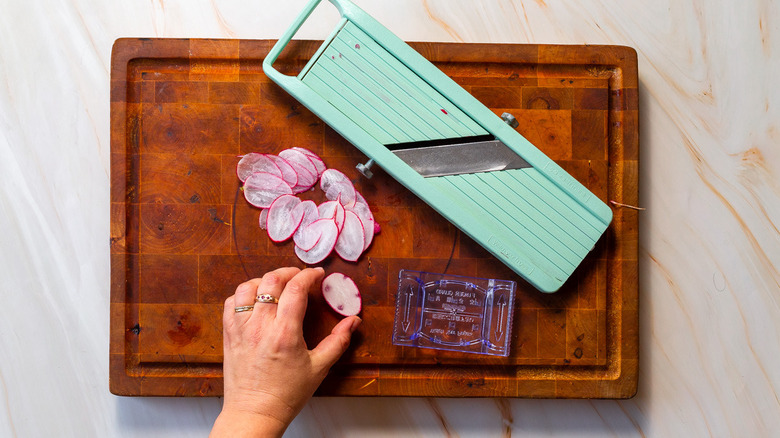 Someone holding a sliced radish near a mandoline on top of a cutting board
