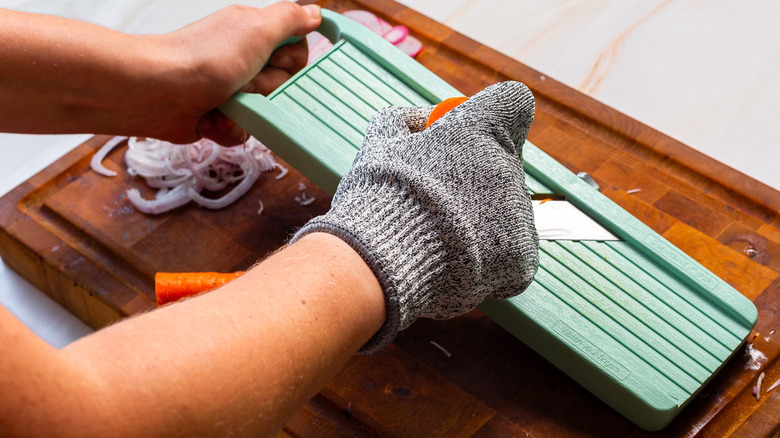 A person wearing a glove on one hand and cutting a carrot on a mandoline placed on a cutting board