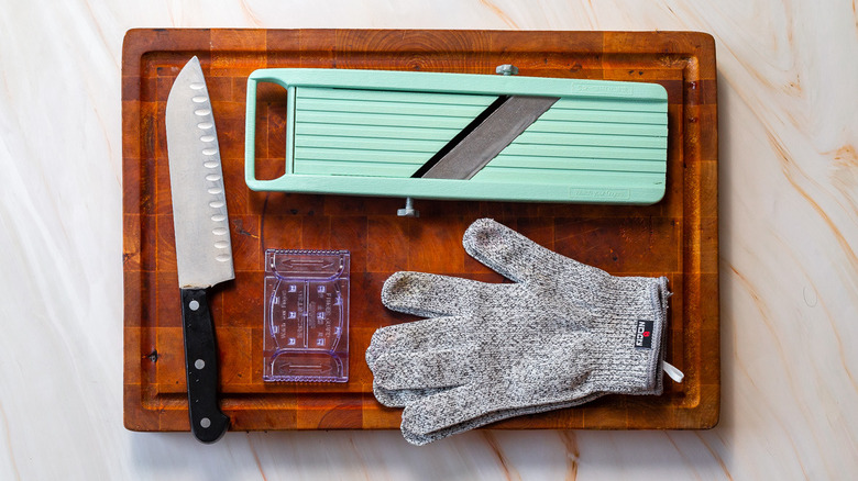 Equipment for using a mandoline slicer, including gloves and a sharp knife, laid out on a wooden cutting board