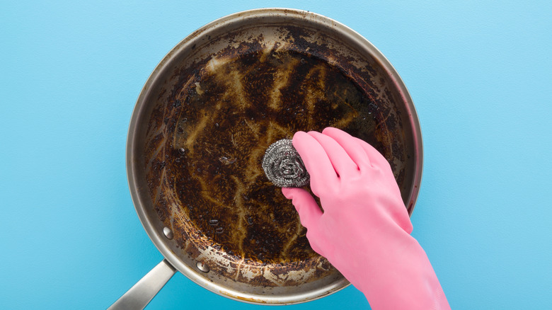 Person scrubbing a dirty stainless steel pan with a steel sponge