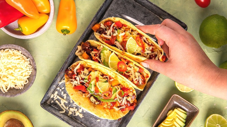 Hand selecting one of three loaded tacos from a metal serving tray