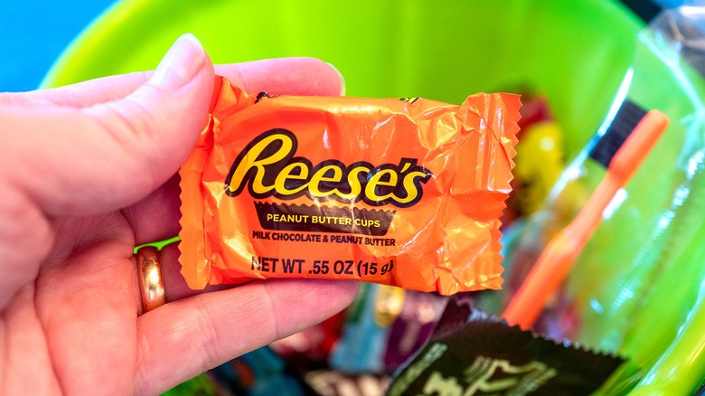 A hand holding a small, individually-wrapped Reese's Peanut Butter Cup over a lime-green bowl filled with candy