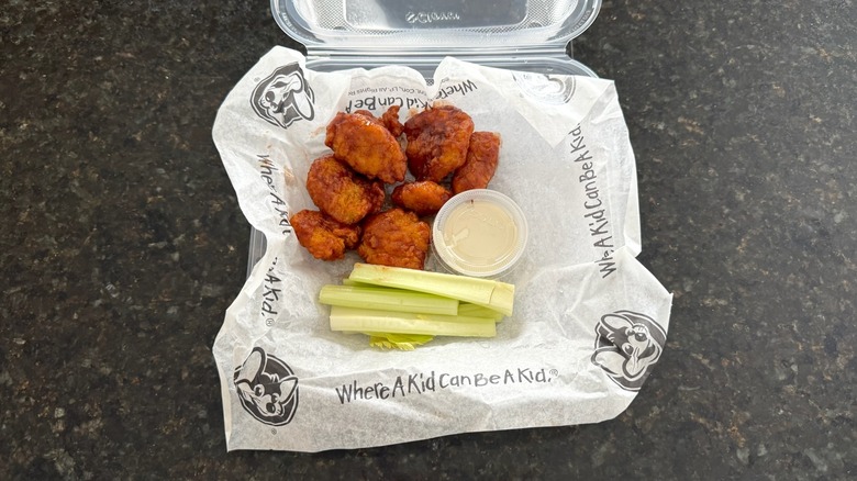 A takeout box of Chuck E. Cheese chicken wings with celery sticks on a black table