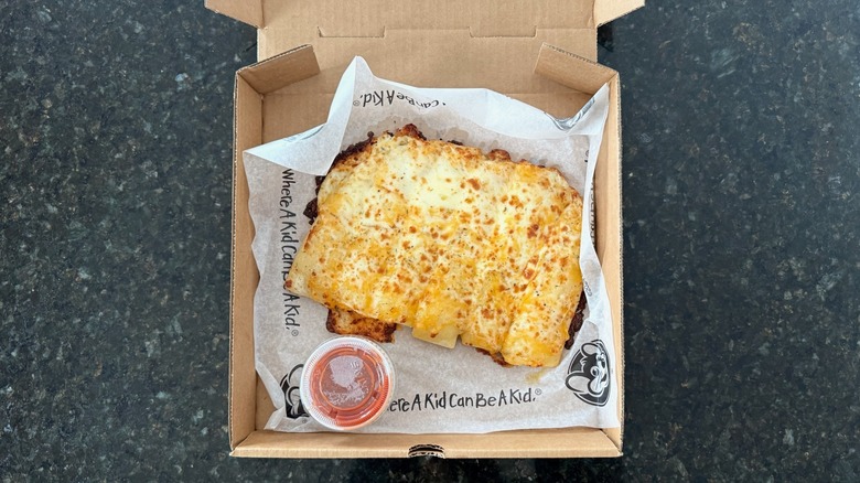 A takeout box of Chuck E. Cheese cheesy bread with a pot of red dipping sauce on the side on a black table