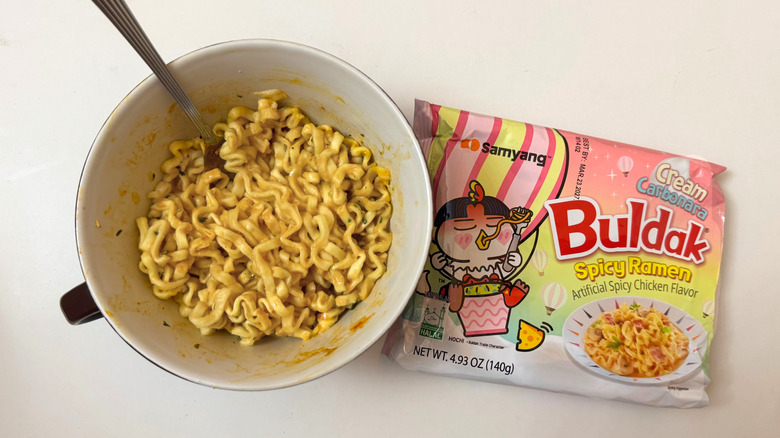 Bowl of Buldak Cream Carbonara noodles next to its original packaging on white surface