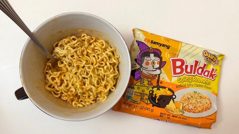 Bowl of Buldak Quattro Cheese noodles next to its original packaging on white surface