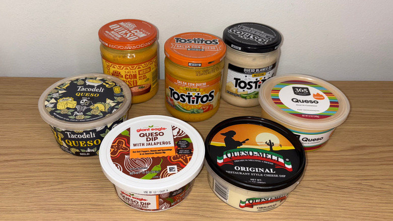 A variety of seven store-bought queso dips in their containers on a wooden table
