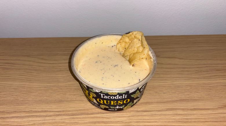 Container of Tacodeli Queso with a tortilla chip in it on a wood table.