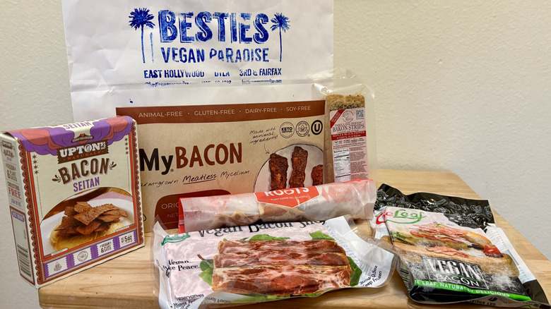 A group of packaged plant-based bacon options in front of a Besties Vegan Paradise shopping bag
