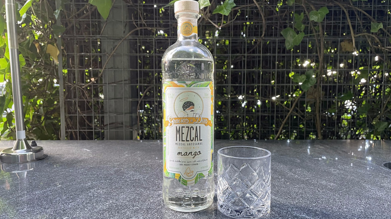 Gracias a Dios mango mezcal bottle and glass tumbler sit on an outdoor counter.