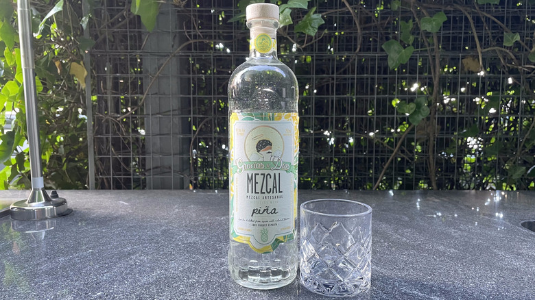 Gracias a Dios piña mezcal bottle and glass tumbler sit on an outdoor counter.