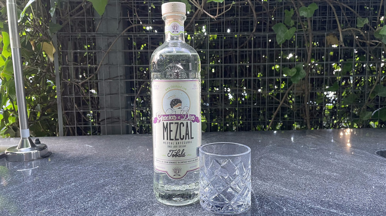 Gracias a Dios Tobala mezcal bottle and glass tumbler sit on an outdoor counter.