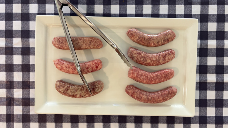 Uncooked brats on white platter with tongs