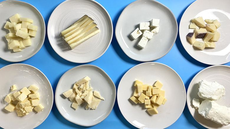 Two rows of white plates with different styles of cut cheeses on a blue surface
