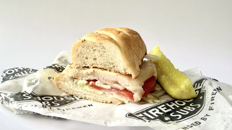 The Smoked Turkey and Provolone from Firehouse with its wrapping and a pickle spear on a white background