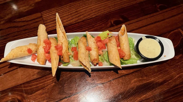 White dish lined with stuffed tortilla rolls and a sauce cup on wooden table.