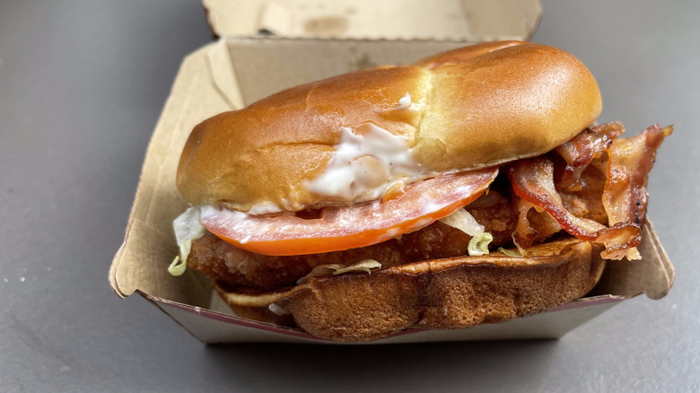 A chicken sandwich with bacon, tomato, and ranch sauce sits in a cardboard container.