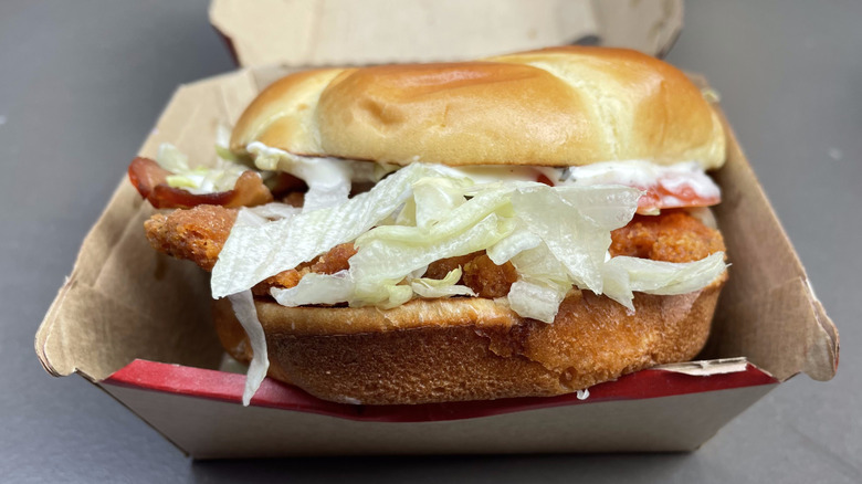An orange chicken patty with lettuce, tomato, bacon, and ranch sauce on a bun in a cardboard container.