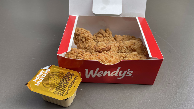 Three crispy chicken tenders in a Wendy's box sit on a table with a honey mustard dipping sauce.