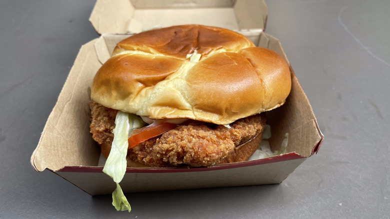 A breaded chicken patty on a bun with tomato, lettuce, and mayo on a bun in a cardboard container sits on a table.