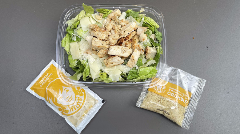 A chicken Caesar salad sits on a table with a packet of Caesar dressing and a packet of Parmesan crumbles.