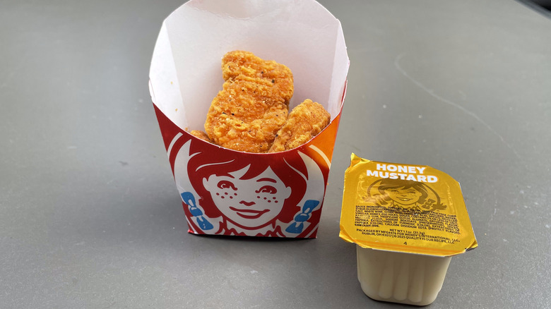 Orange colored chicken nuggets sit in a Wendy's french fry container on a table with a honey mustard cup.