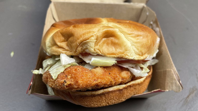 An orange chicken breast with tomato, lettuce, and mayo on a bun in a cardboard container.