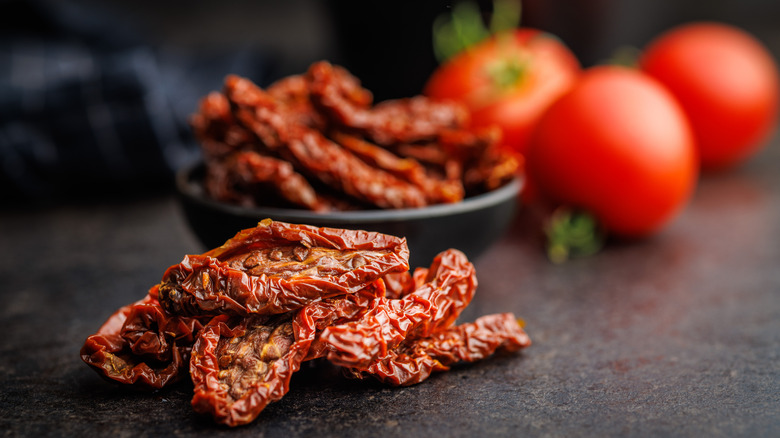 A pile of sun-dried tomatoes on a black counter with whole tomatoes in the background