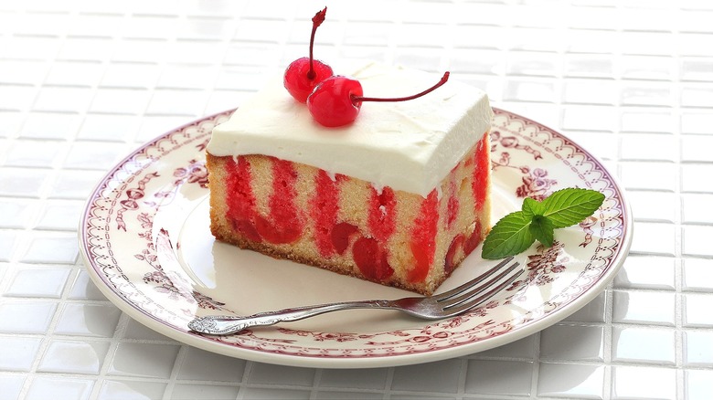 Piece of cherry poke cake on a cream colored plate with burgundy trim