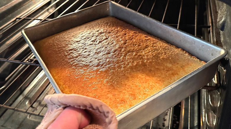 Hand holding an oven mitt pulling a rectangular vanilla cake out of an oven