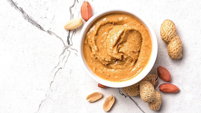 Small white bowl with natural peanut butter surrounded by peanuts