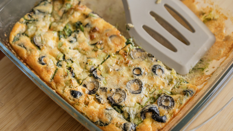 Part of an egg casserole with spinach and olives sits in a clear glass baking dish with a spatula on a wooden table.