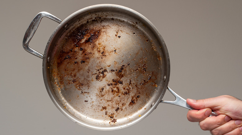 Hand holding the handle of a dirty stainless-steel pan with stuck on food.