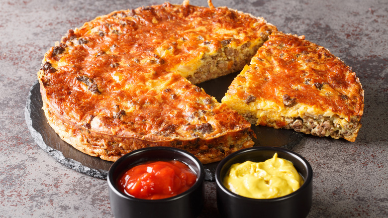 Cheeseburger pie with two slices cut out on a gray slate with two cups of ketchup and mustard