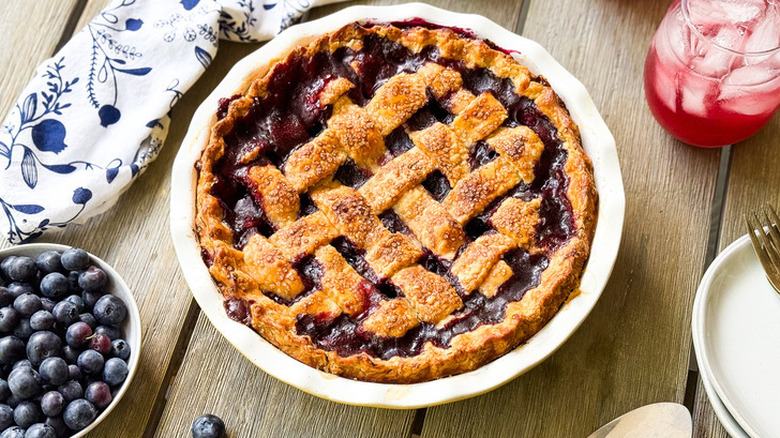 A freshly baked blueberry pie with a lattice crust aside a bowl of blueberries and a glass of juice.